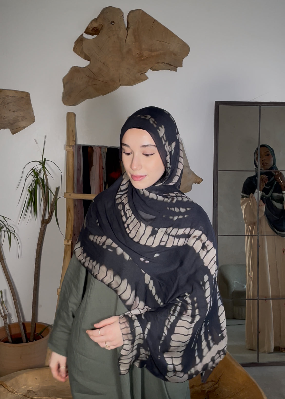 Woman wearing a patterned hijab in a room with plants and a mirror.